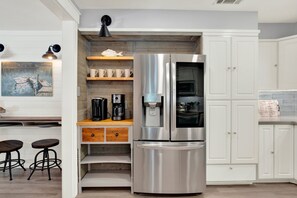 A modern kitchen with white cabinetry, a stainless steel refrigerator, a coffee maker, and a wooden shelf with cups. - Private Heated Pool, Family Beach Getaway (Tybee Island)