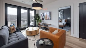 Un salon moderne avec un canapé, une table basse et un téléviseur. Il y a une grande fenêtre et une porte qui donne sur une autre pièce.