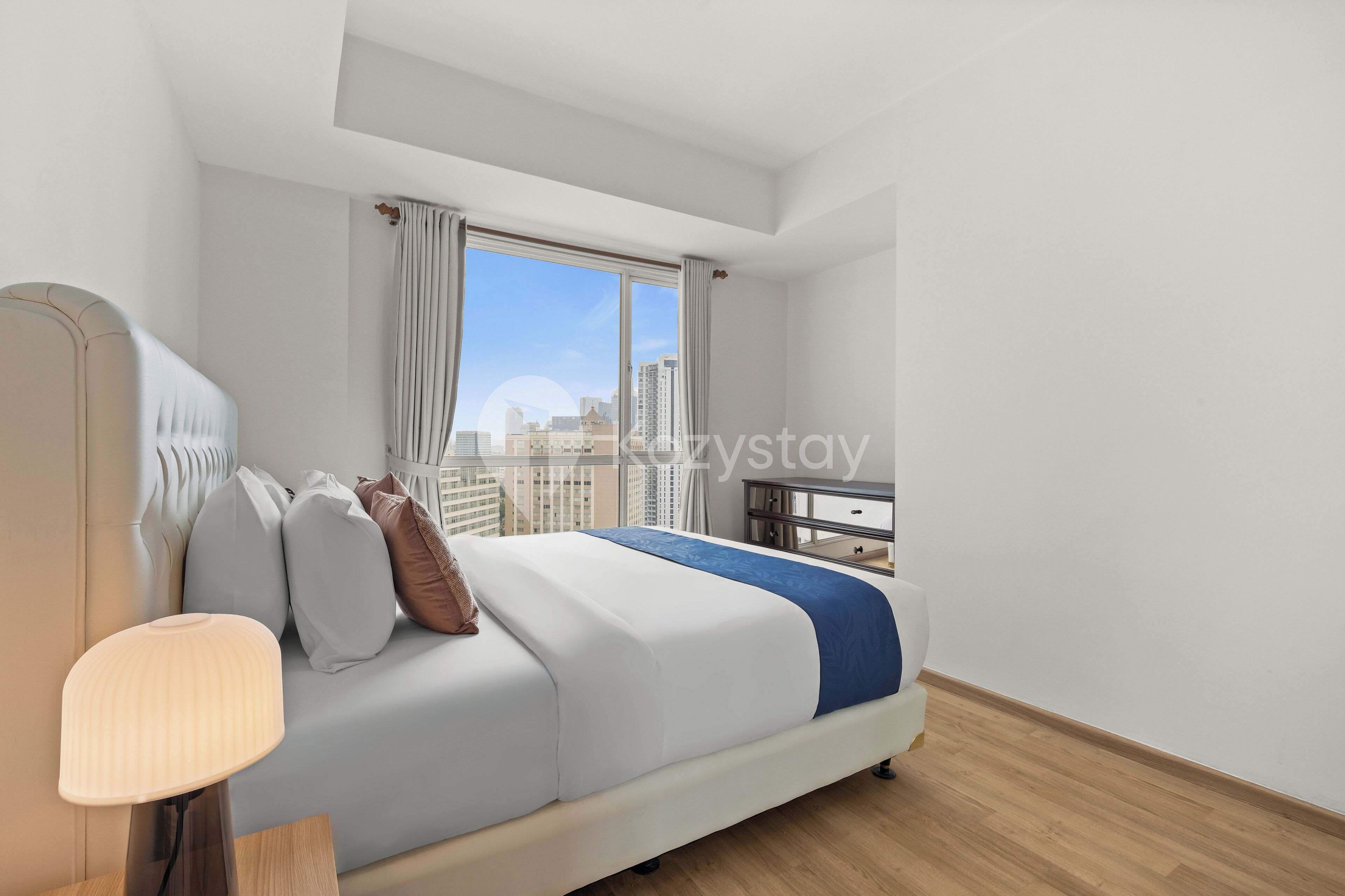 A modern hotel room with a large bed, a bedside table, a lamp, and a view of the city skyline through a large window.