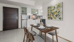 A modern kitchen with white cabinets, a dining table set with white plates and silverware, and a large abstract painting on the wall. - Serenya by Kozystay - Kuningan (Jakarta)