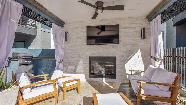 A patio area with a ceiling fan, a flat-screen TV mounted on the wall, a fireplace, and wooden furniture. - Scottsdale 2BR/2BA • Pool • Gym • Free Parking (Phoenix)