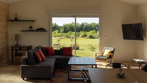 A spacious living room with a large sectional sofa, a wooden dining table, and a view of a garden through a sliding glass door. - Renovated farmhouse with swimming pool, pétanque and country views (Coufouleux)