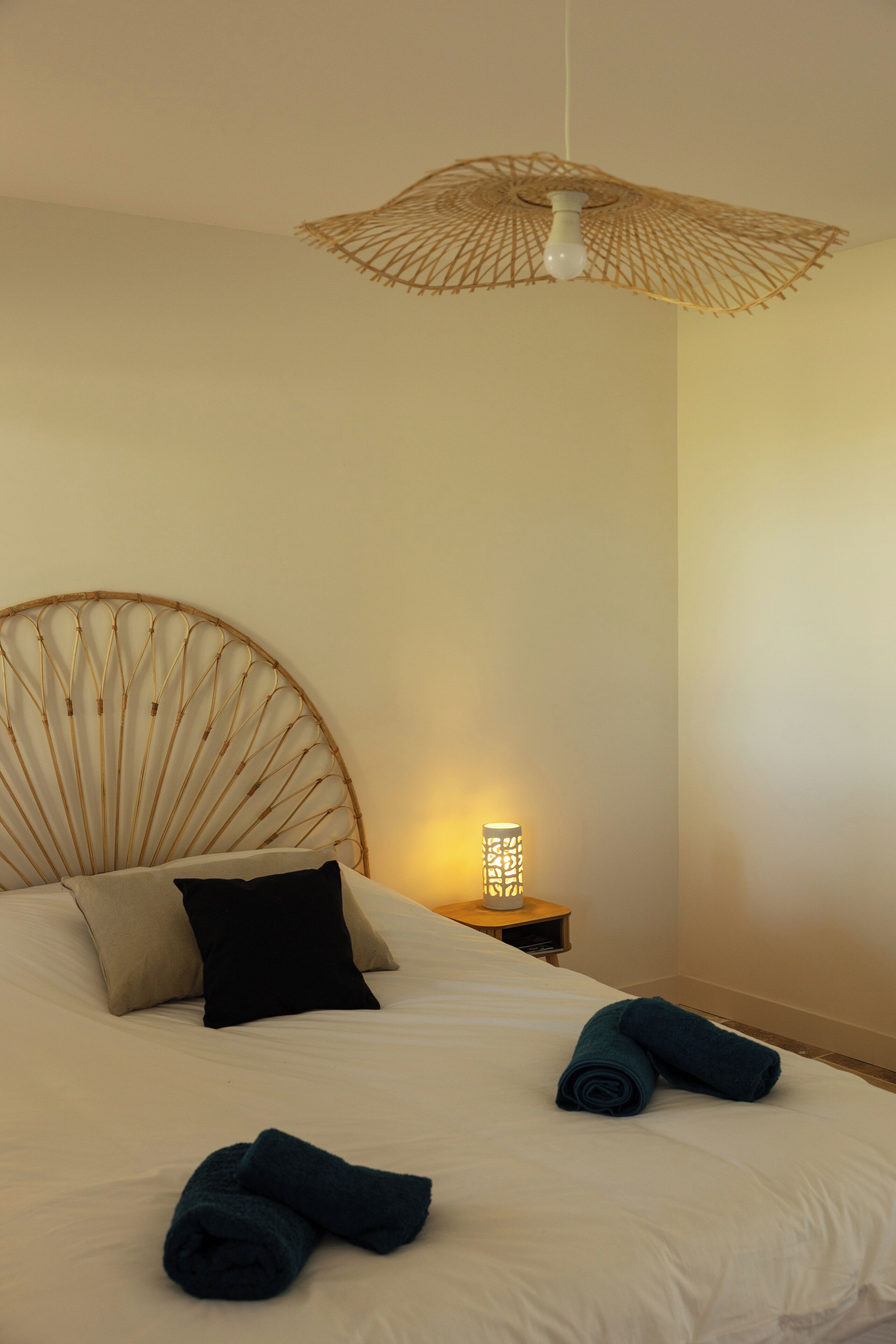 Ein ordentlich bezogenes Bett mit zwei aufgerollten Handtüchern, einer Nachttischlampe und einem gewobenen Kopfteil.