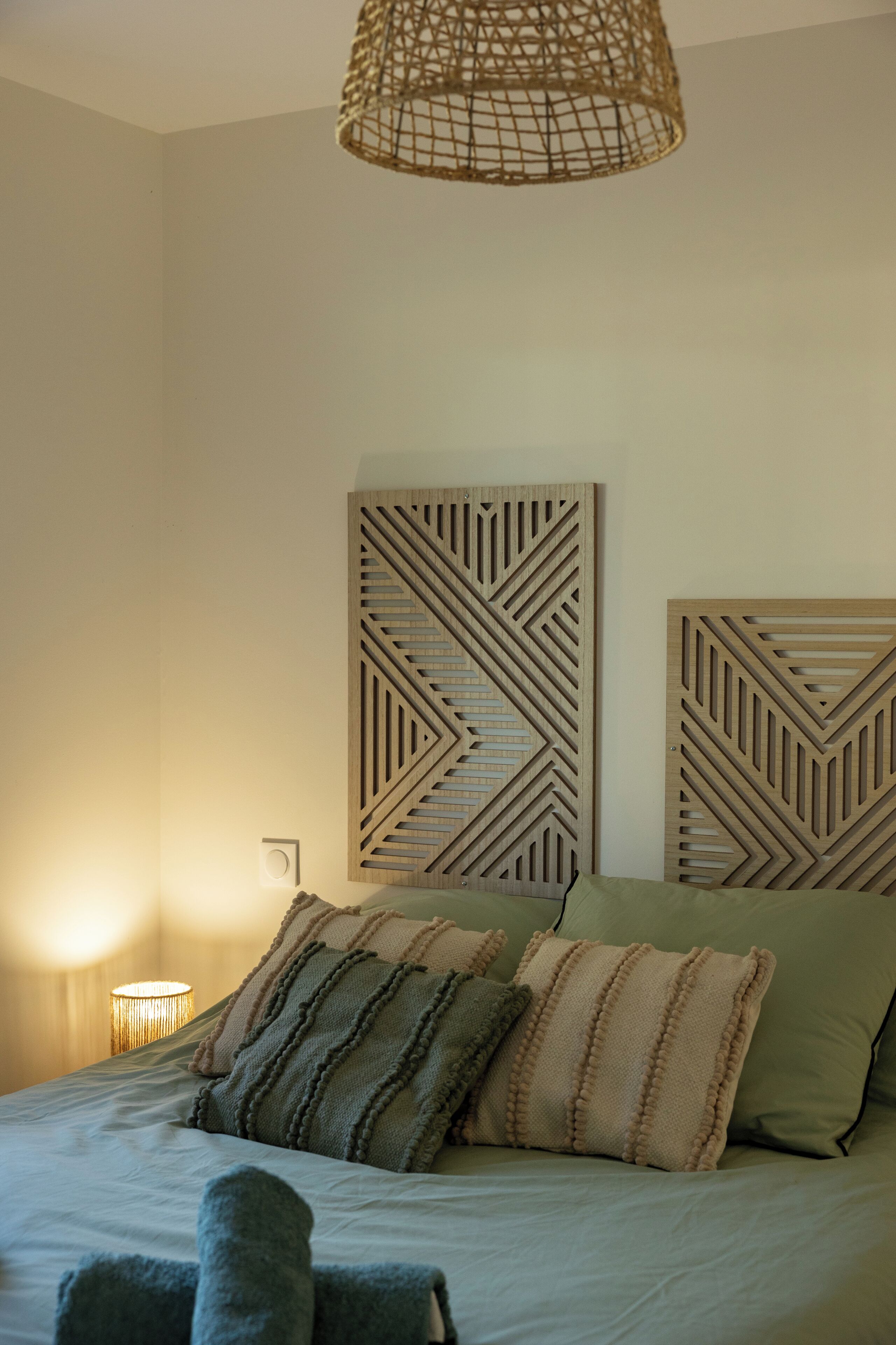 Un dormitorio con cama, almohadas, una lámpara fijada a la pared y un panel de madera decorativo.