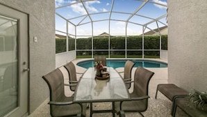 A covered patio with a glass table and chairs, a pool, and a view of a green landscape.