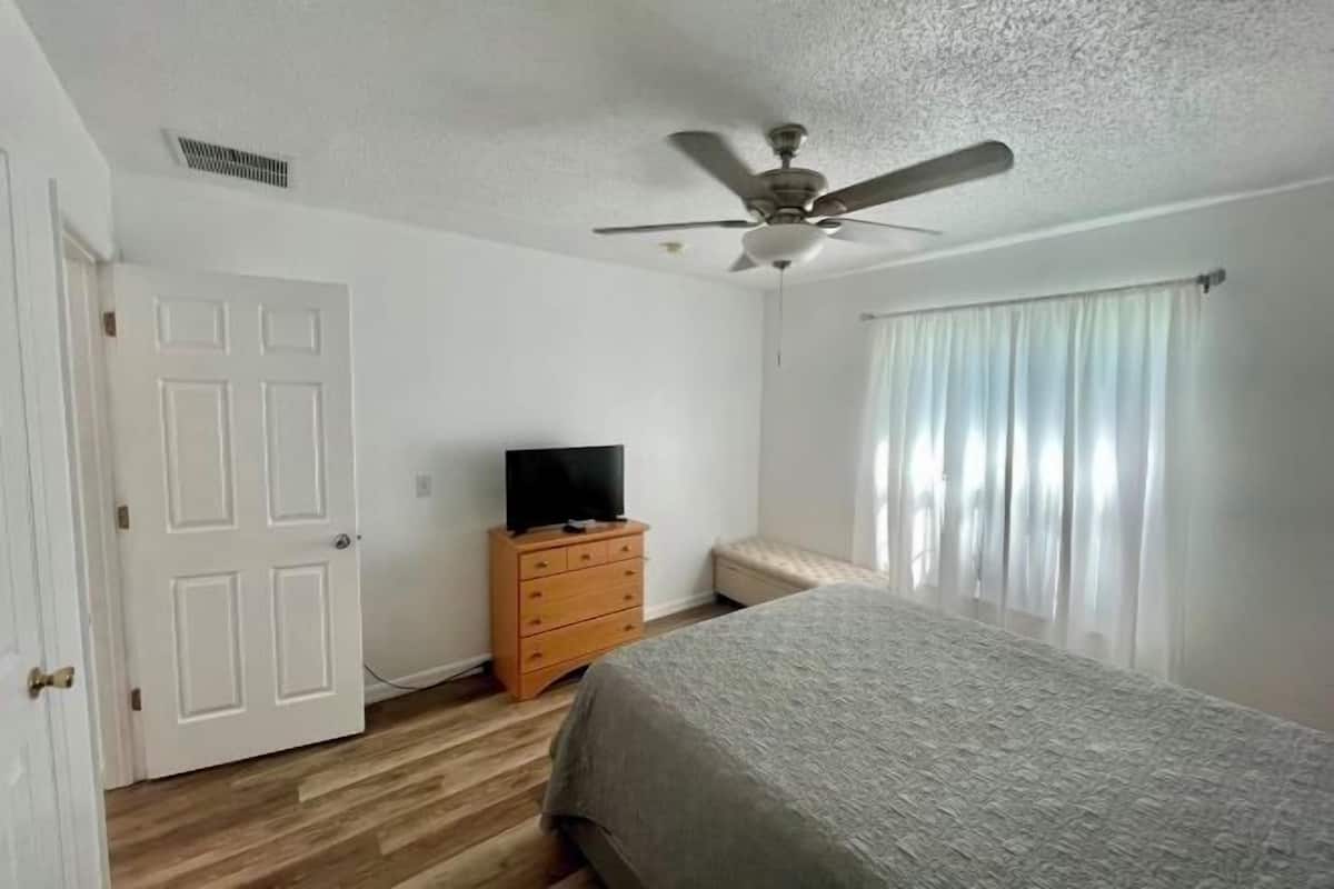 Une chambre à coucher comprenant un lit, un ventilateur de plafond, une commode, un téléviseur et une fenêtre avec des rideaux.