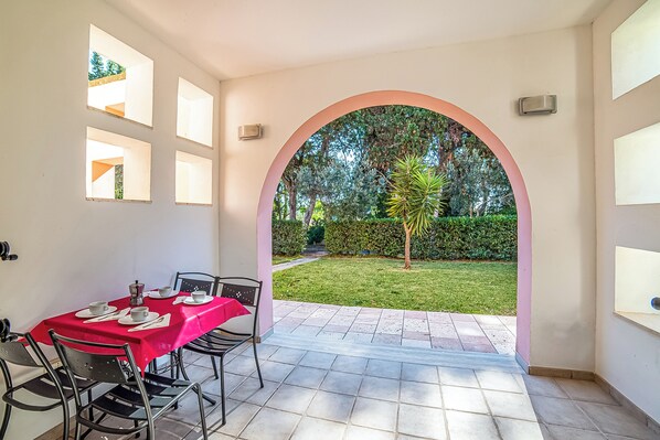 Un'area pranzo all'aperto con tovaglia rossa, sedie e vista su un giardino attraverso un arco.