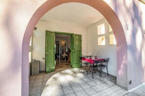 Un arco d'ingresso che dà su una sala da pranzo con tavolo e sedie, un piccolo cestino e una porta con persiane verdi.
