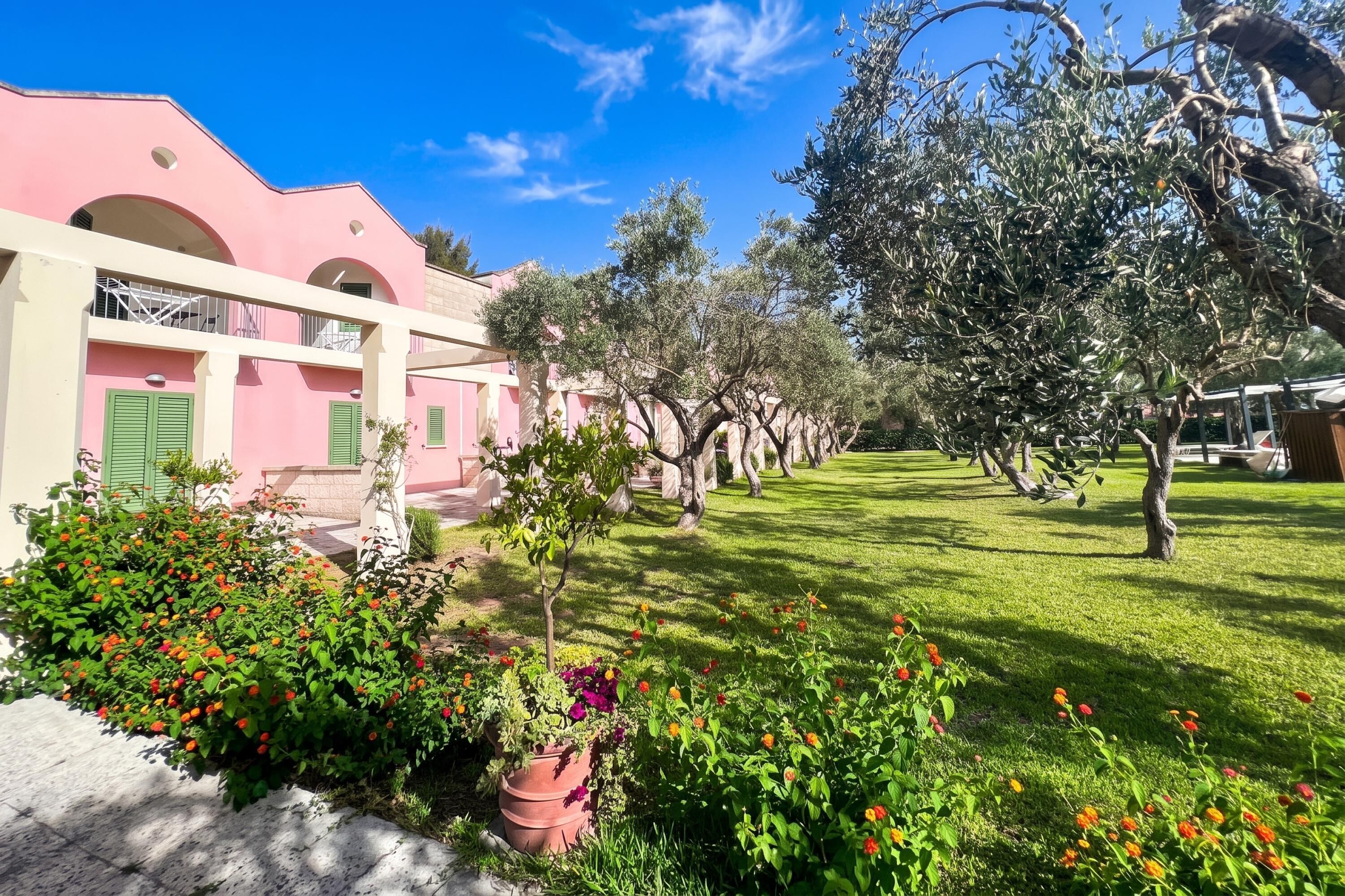 Ein pinkfarbenes Gebäude mit Balkon, umgeben von einem gepflegten Garten mit verschiedenen Pflanzen und Blumen.
