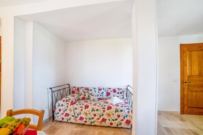 Ein kleiner, abgeschlossener Raum mit einem Bett, einer Holztür und einem Stuhl mit Obstschale.