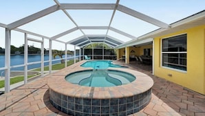 A covered outdoor swimming pool area with a hot tub, surrounded by a glass enclosure, and a view of a lake. - Casa de La Vista – Pool & Spa with lakefront view (Cape Coral)