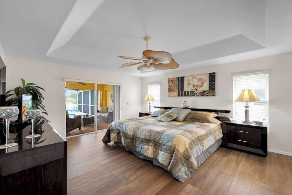 A bedroom with a bed, a ceiling fan, a nightstand, a lamp, and a large window.
