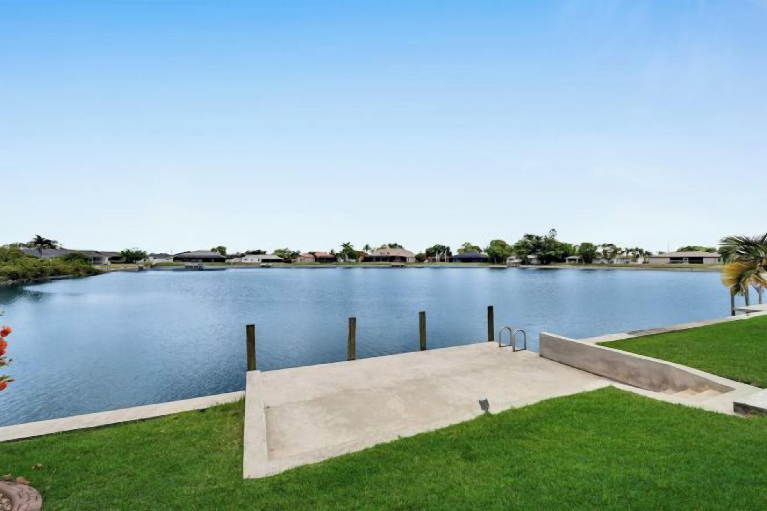 A calm lake with a concrete dock, surrounded by a grassy area and distant buildings.