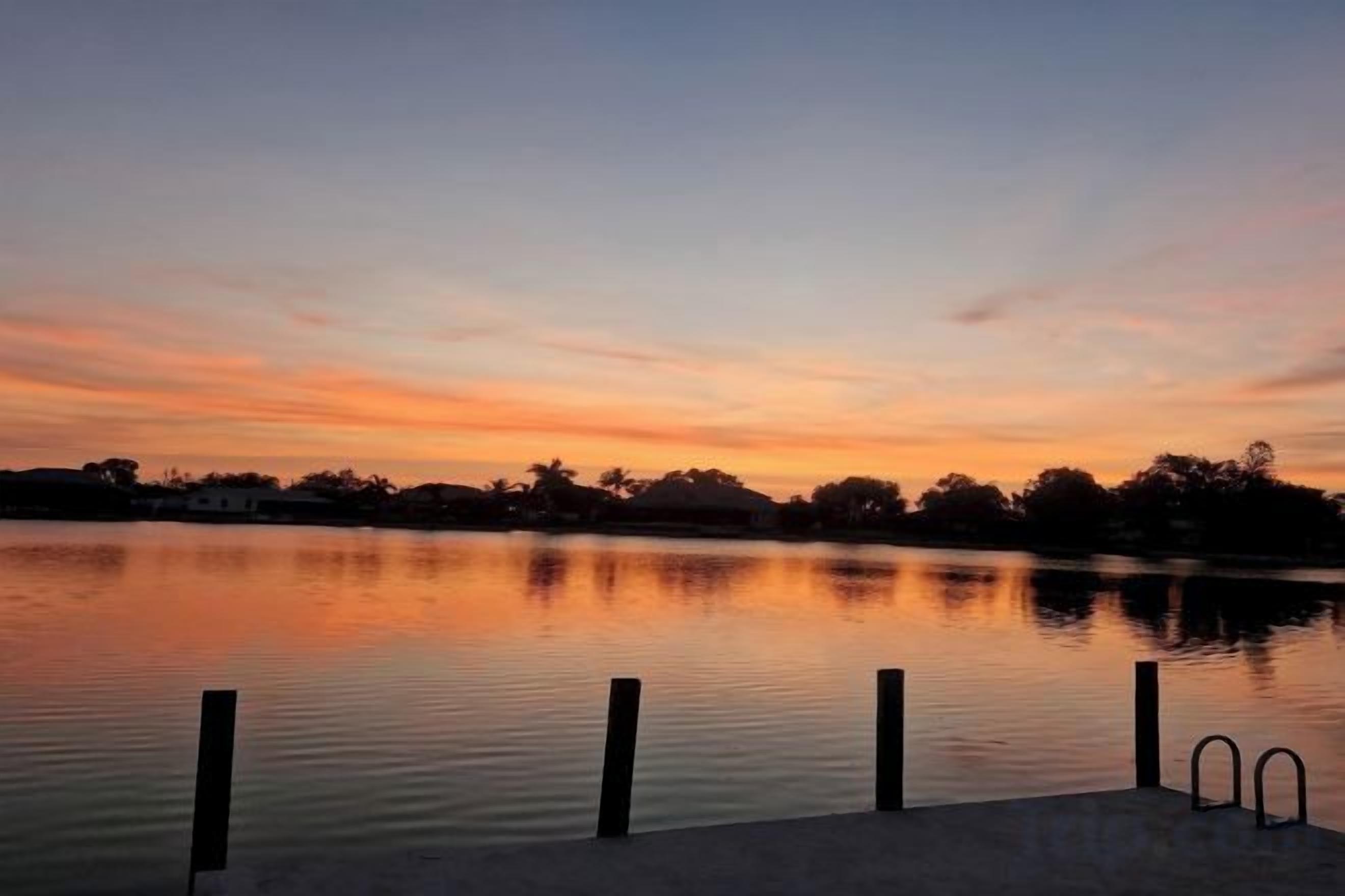 A calm body of water reflecting a sunset sky, a dock with a ladder, and a distant tree line.