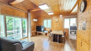A cabin interior with a sofa, dining table, chairs, a television, and a clock on the wall. - Zain Village pension (Seogwipo)
