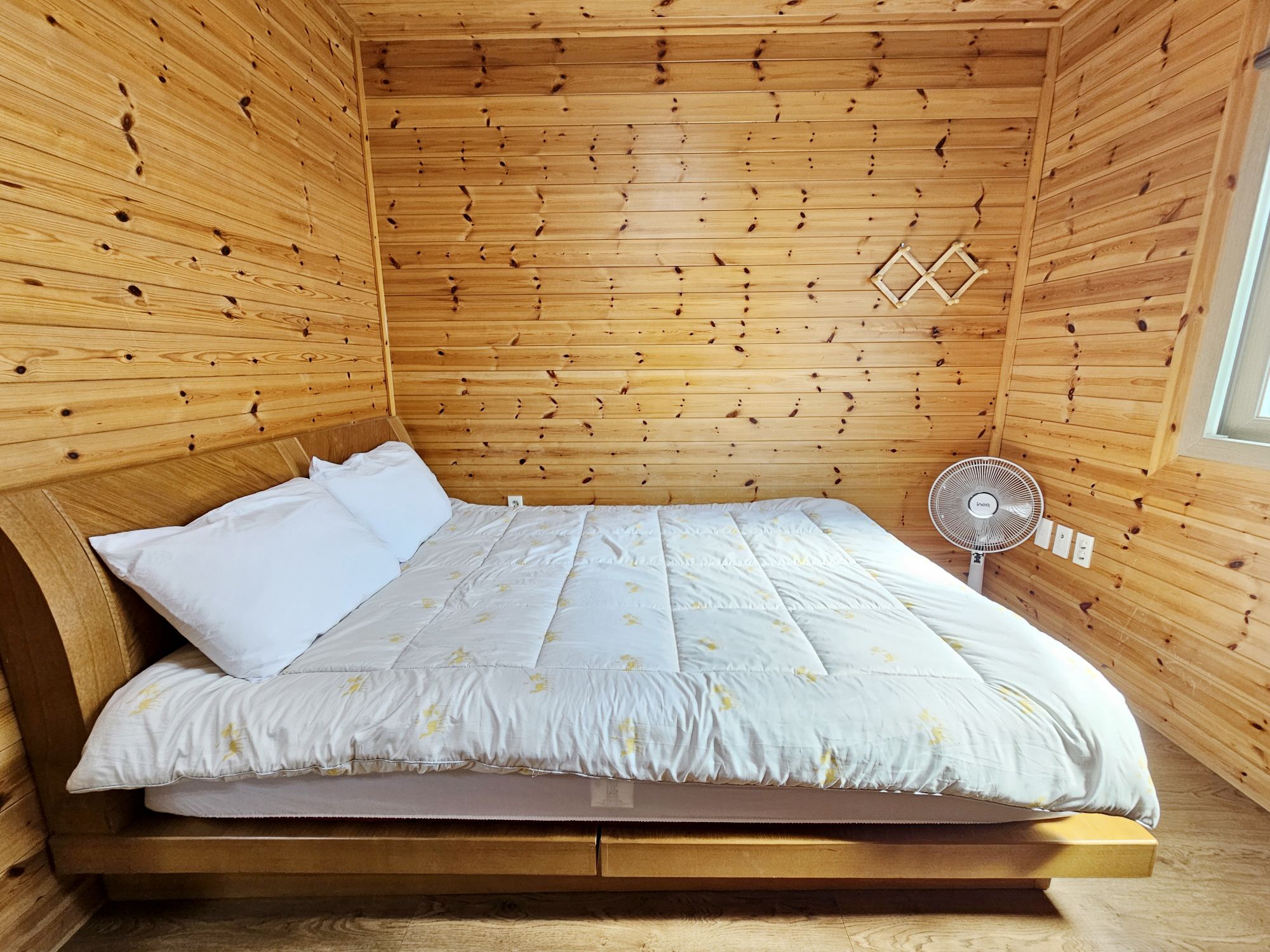 A wooden cabin room with a bed, a fan, and a window.