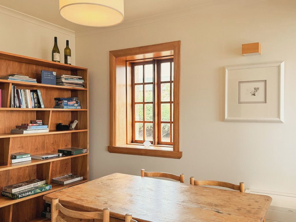 A room with a wooden table, chairs, a bookshelf, a window, and a framed picture on the wall.