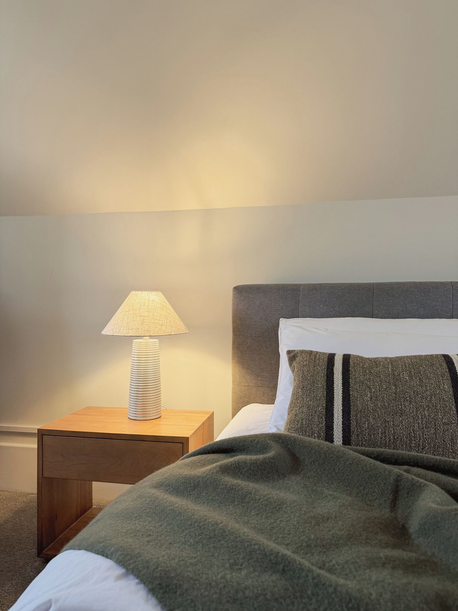 A neatly made bed with a grey headboard and a patterned pillow, a bedside lamp on a wooden nightstand, and a carpeted floor.