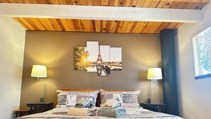 A bedroom with a wooden ceiling, a bed with patterned bedding, two bedside lamps, and a wall with a framed picture of the Eiffel Tower.
