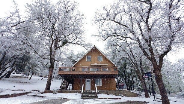 A two-story wooden house with a balcony, surrounded by snow-covered trees.