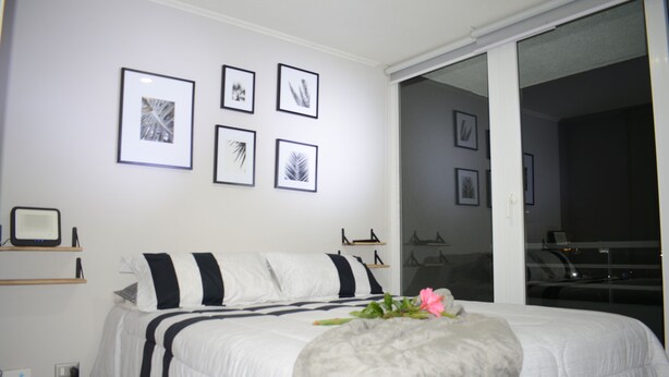 A bedroom with a bed, a bedside table, a wall-mounted shelf, and a sliding glass door leading to a balcony. - Apartamento 2 dormitorios, cerca de lo mas destacado de la ciudad y aeropuerto (Santiago)