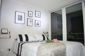 A bedroom with a bed, a bedside table, a wall-mounted shelf, and a sliding glass door leading to a balcony. - Apartamento 2 dormitorios, cerca de lo mas destacado de la ciudad y aeropuerto (Santiago)