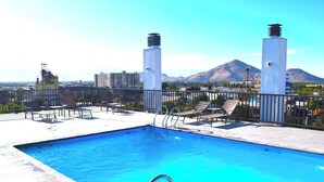 A rooftop pool with clear blue water, surrounded by lounge chairs and a cityscape with mountains in the background. - Apartamento 2 dormitorios, cerca de lo mas destacado de la ciudad y aeropuerto (Santiago)