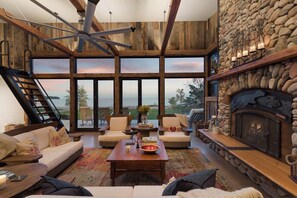 A spacious living room with a stone fireplace, large windows, and a wooden ceiling fan. - Stone and Cedar Lodge (LaPointe)