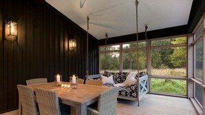 Une terrasse en bois avec une table et des chaises, un banc rembourré agrémenté de coussins à motifs et une grande fenêtre donnant sur un paysage verdoyant.