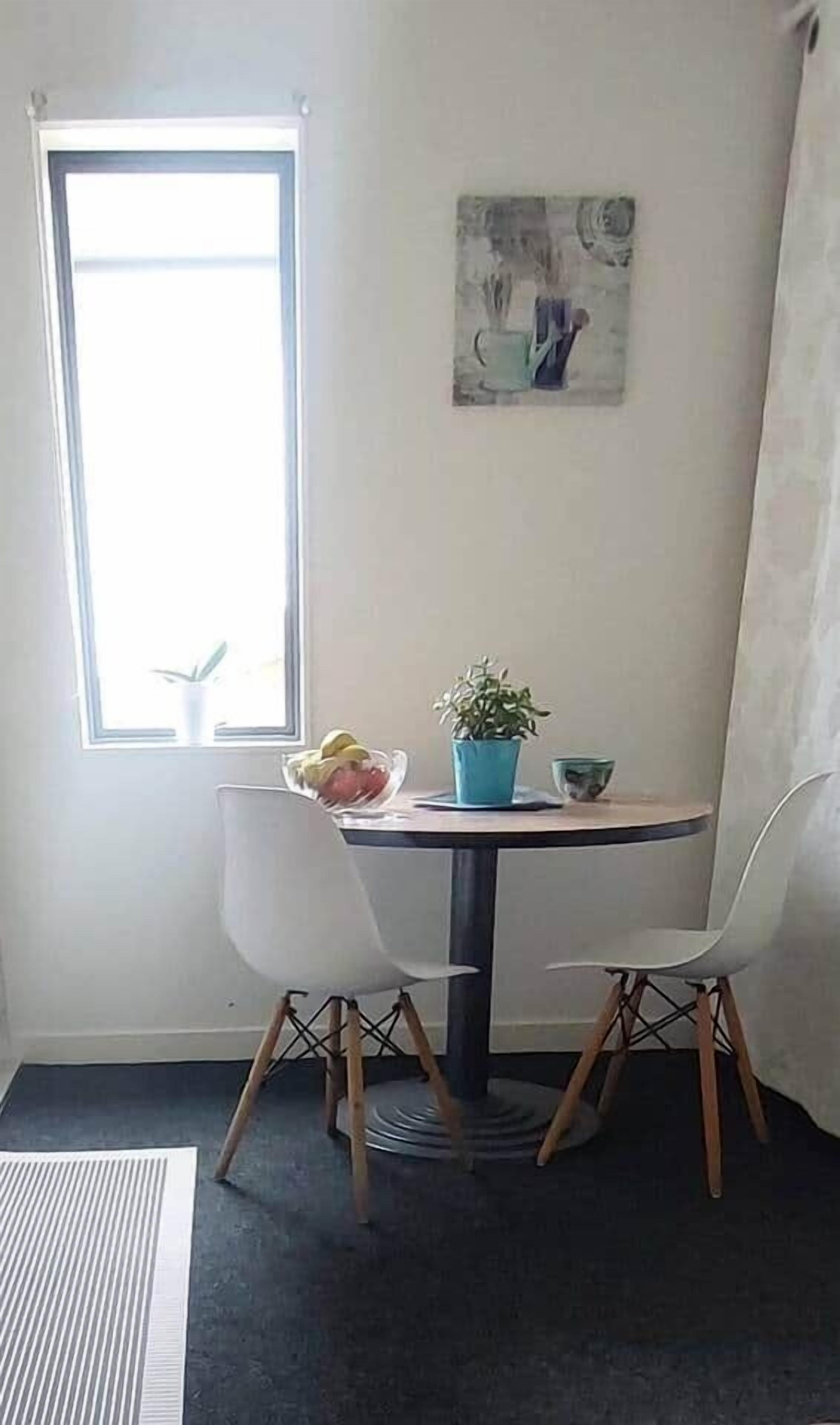 Pieni ruokailutila, jossa on pyöreä pöytä, kaksi valkoista tuolia, ruukkukasvi, hedelmäkulho ja ikkuna, josta on näkymä.