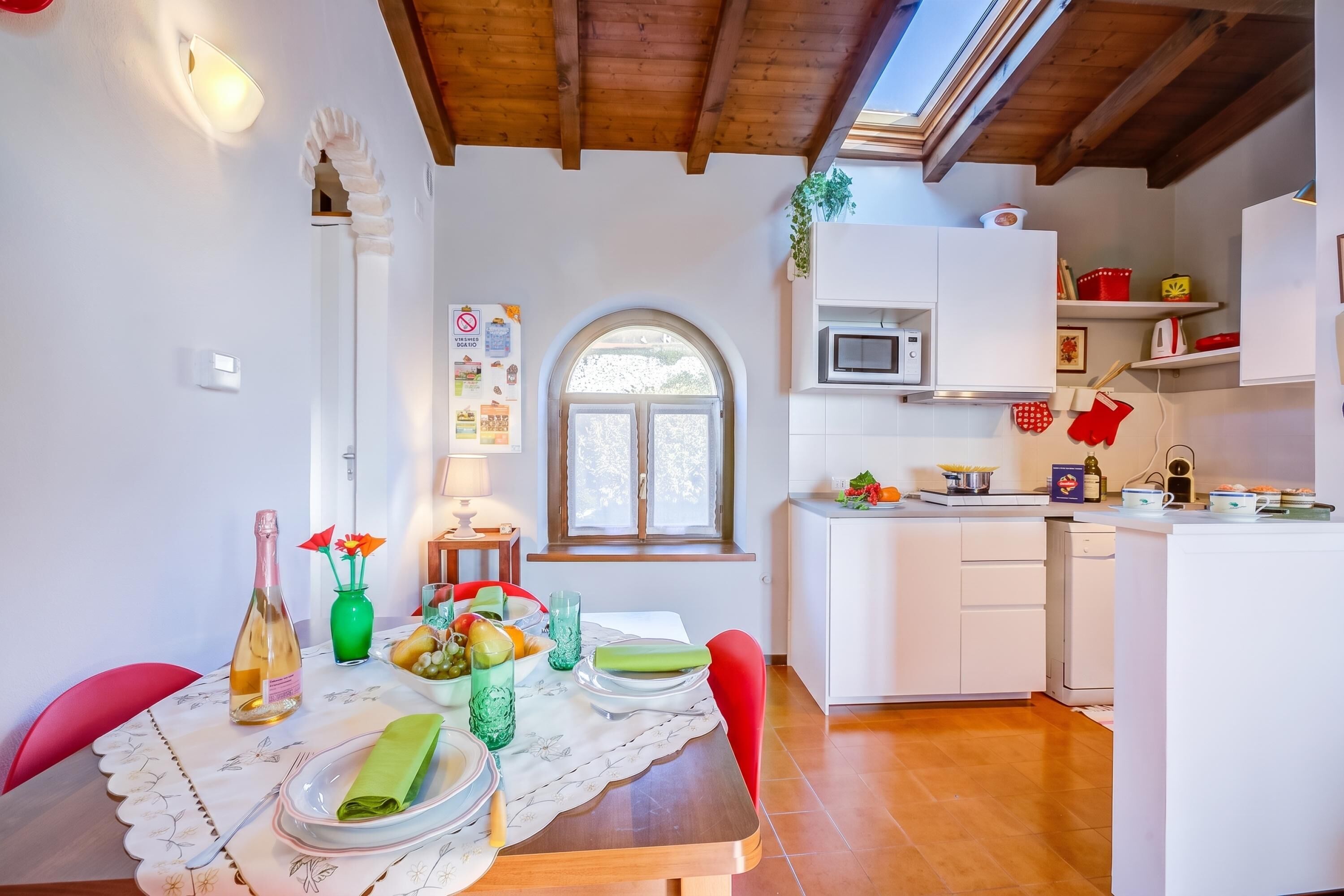 A dining area with a table set for two, a kitchen with white cabinets, and a window with a view of greenery.