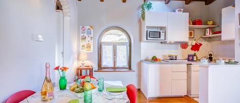 A dining area with a table set for two, a kitchen with white cabinets, and a window with a view of greenery.