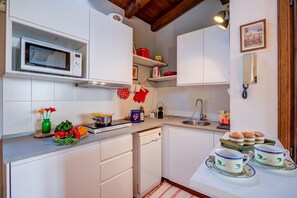 A compact kitchen with white cabinets, a microwave, a sink, and a dishwasher. There are decorative items like a vase with flowers and a plate of vegetables on the counter. - Le Fontane With View Lake (Gardone Riviera)