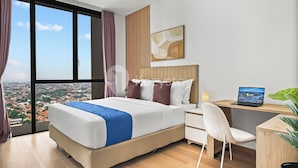 A hotel room with a large bed, a desk with a laptop, a chair, and a view of a cityscape through large windows. - Osvara by Kozystay - Cilandak (Jakarta)