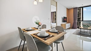 A modern dining area with a wooden table, grey chairs, and a vase of flowers. A television is mounted on the wall, and a large window offers a view of the city. - Osvara by Kozystay - Cilandak (Jakarta)