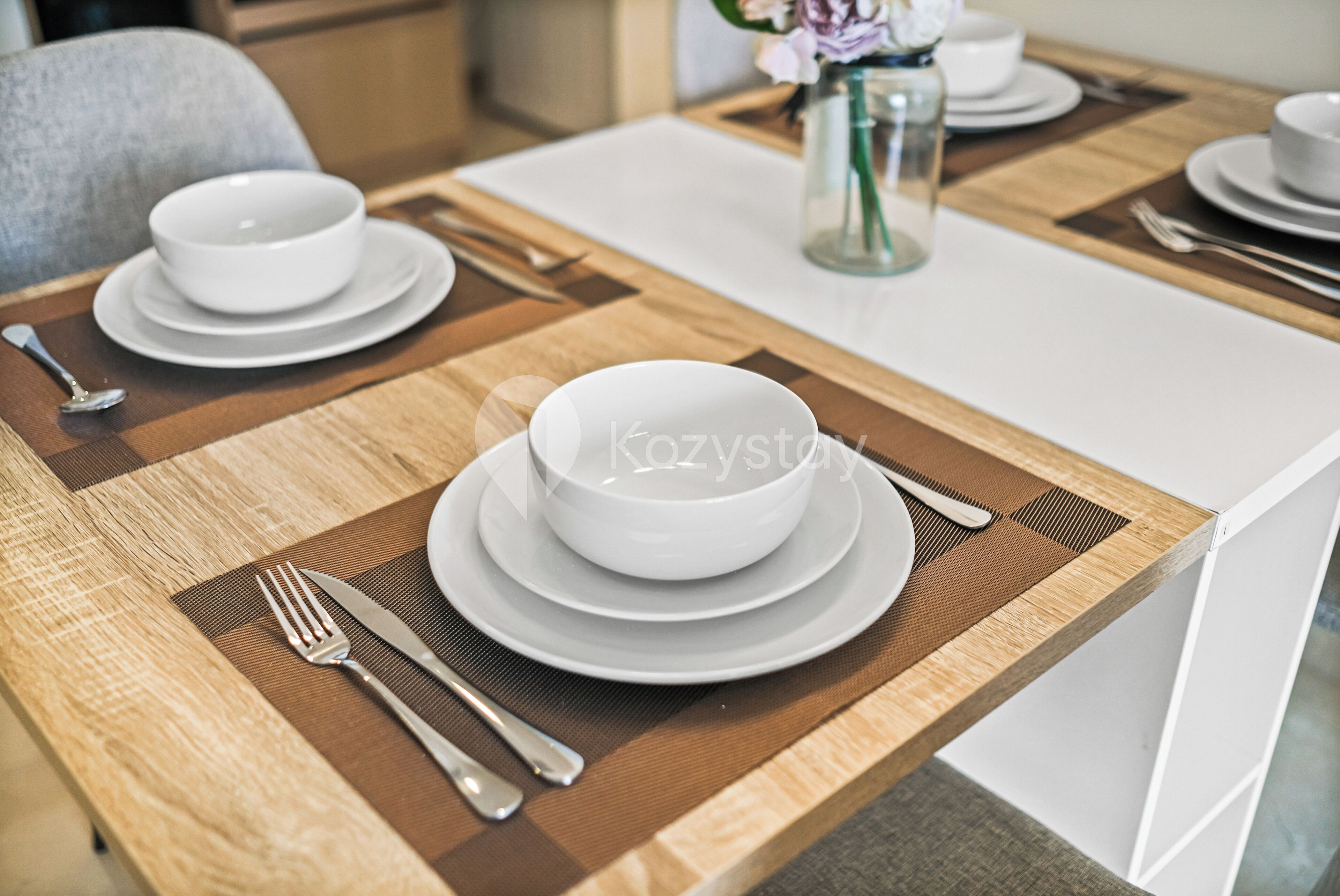 A dining table set with white plates, bowls, and cutlery, with a placemat and a vase of flowers in the background.