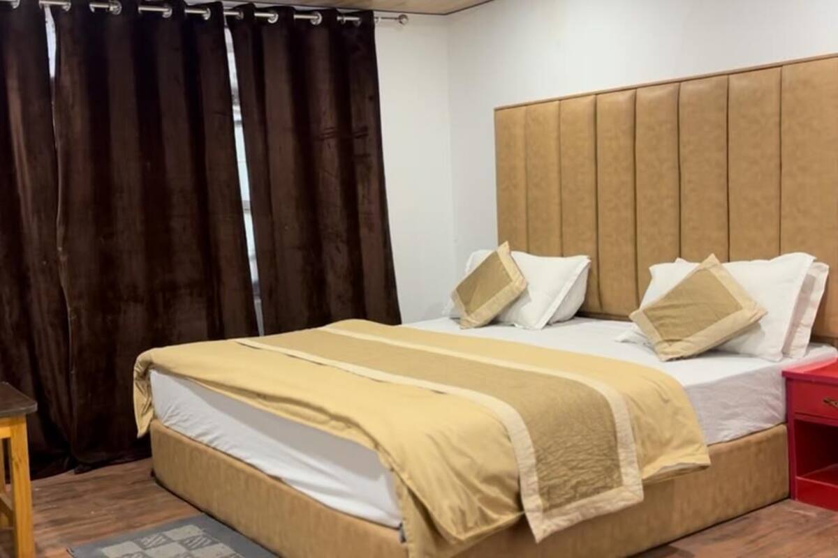 A hotel room with a bed, a wooden nightstand, a red side table, and a dark curtain.