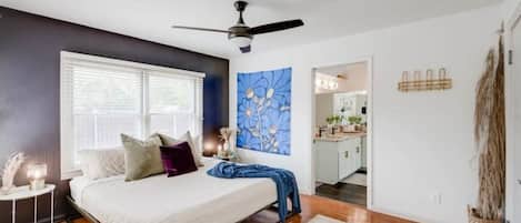 A bedroom with a bed, a ceiling fan, a window with blinds, a painting on the wall, and a view into the kitchen.