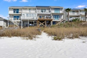 A beachfront hotel with multiple balconies, a sandy path leading to the entrance, and a wooden deck area.