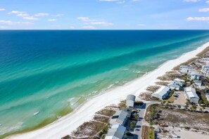 An aerial view of a coastal area with a beach, clear blue waters, and a cluster of buildings with flat roofs.