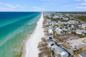 An aerial view of a coastal area with a beach, residential buildings, and a road.