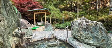 A traditional Japanese onsen with a wooden bathhouse, stone steps, and a natural forest backdrop.