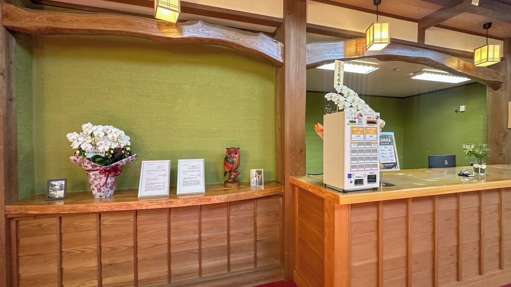 A reception desk with a green wall, wooden paneling, a vase of white flowers, framed certificates, and a small owl figurine.