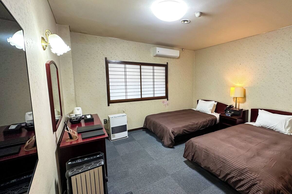 Habitación de hotel con dos camas, un escritorio con silla, un espejo, un equipo de aire acondicionado y una ventana con persianas.