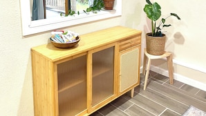 Una consola de madera con un espejo encima, una planta en maceta sobre un taburete, y una ventana con cortinas.