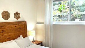 Una cama bien tendida con sábanas blancas y cabecera de madera, una mesita de noche con lámpara, y una ventana con cortinas.
