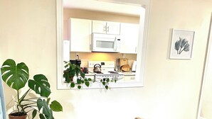 Una pequeña kitchenette con gabinetes blancos, un microondas y una estufa. Hay un banco de madera con un zapatero debajo y una planta en maceta a la izquierda.