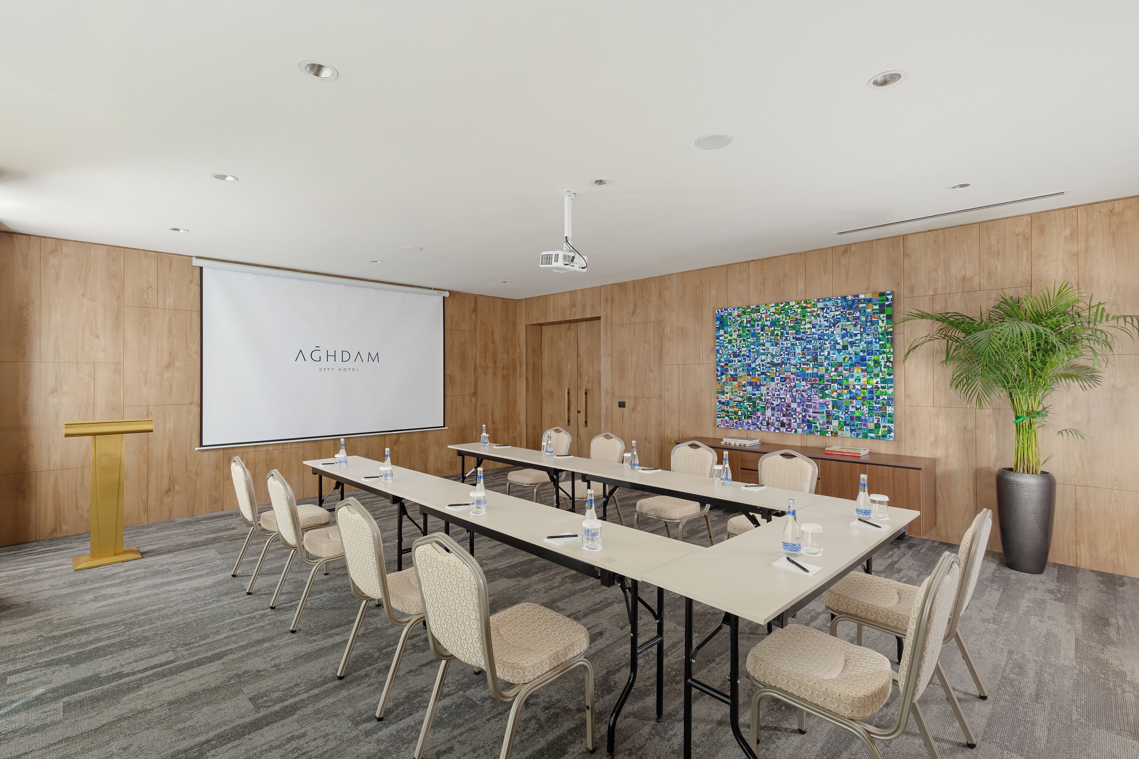 A conference room with a long table, chairs, a podium, a projector screen, and a colorful abstract painting on the wall.