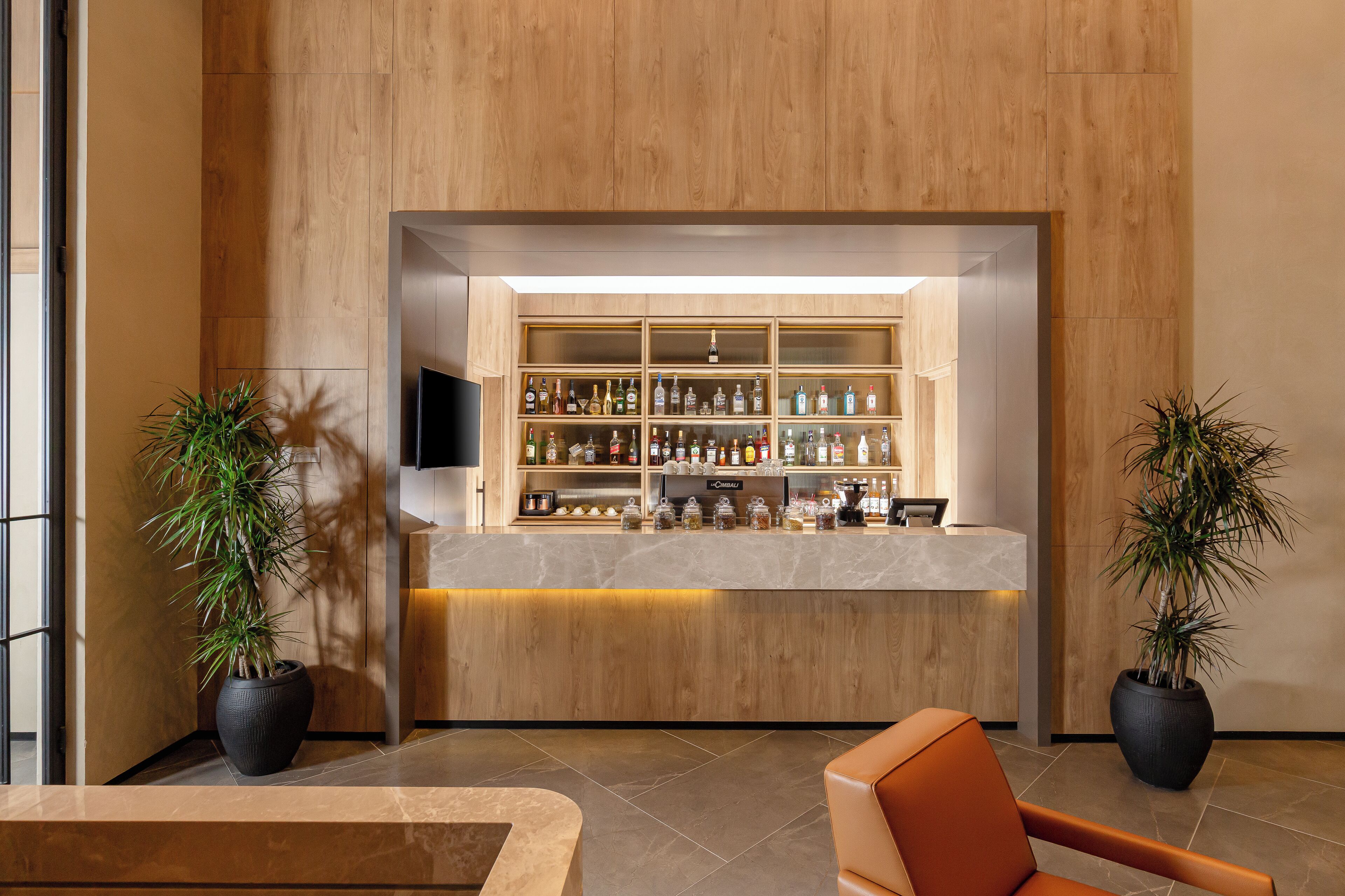 A reception area with a wooden reception desk, a television, and a bar stocked with various bottles. There are two potted plants on either side of the desk.