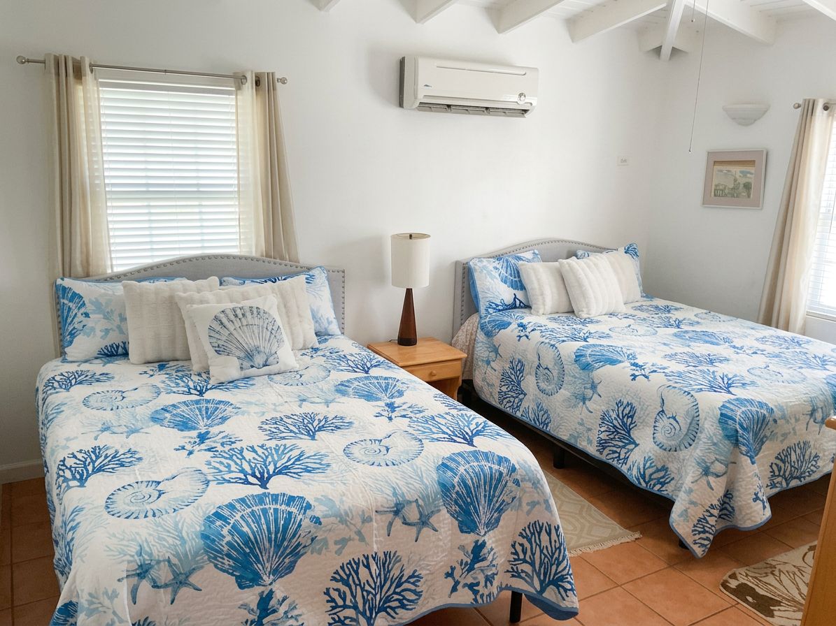 A bedroom with two beds, each with a blue and white floral bedspread, a bedside table with a lamp, and an air conditioning unit on the wall.